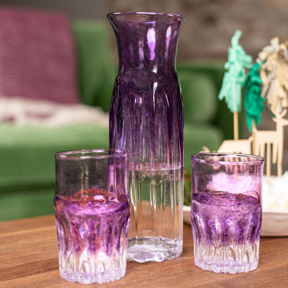 Carafe and two glasses with clear bottom and purple glass top, half filled with a clear liquid, on a wood table with a green couch in the background. 