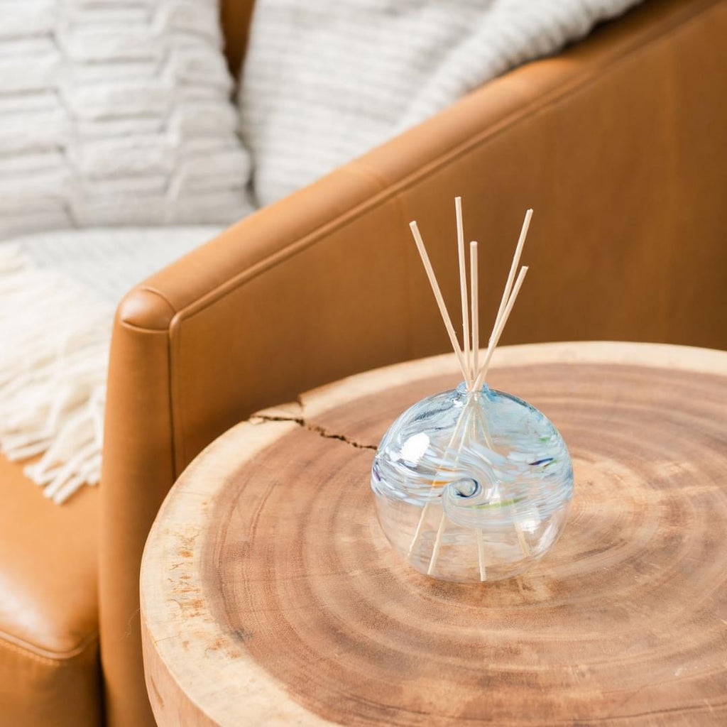 A clear glass orb with a flat bottom with white, blue, red and lime and glass swirled on the top half. Five reeds are in a hole in the top of the orb. Displayed on a wooden table beside a lamp with a book and couch in the background.