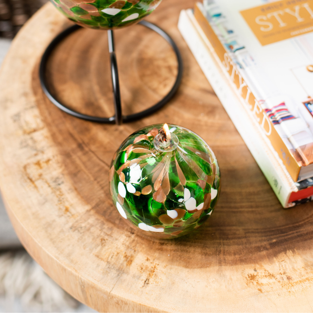 Round glass ball with flat base and wick with green, brown glitter and opaque white glass mosaiced around the entire surface on a wooden side table with a stack of books to the right.