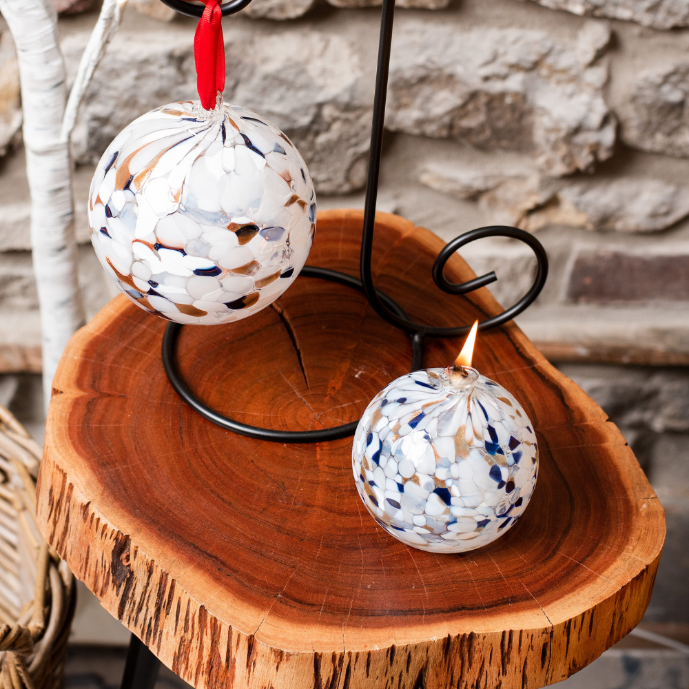 Round glass ball with flat base and wick with opaque navy glitter, opaque copper glitter and opaque white glass mosaiced around the entire surface on a wooden side table with a Round glass ball with opaque navy glitter, opaque copper glitter and opaque white glass mosaiced around the entire surface on a black ornament holder. Stone wall in the background.