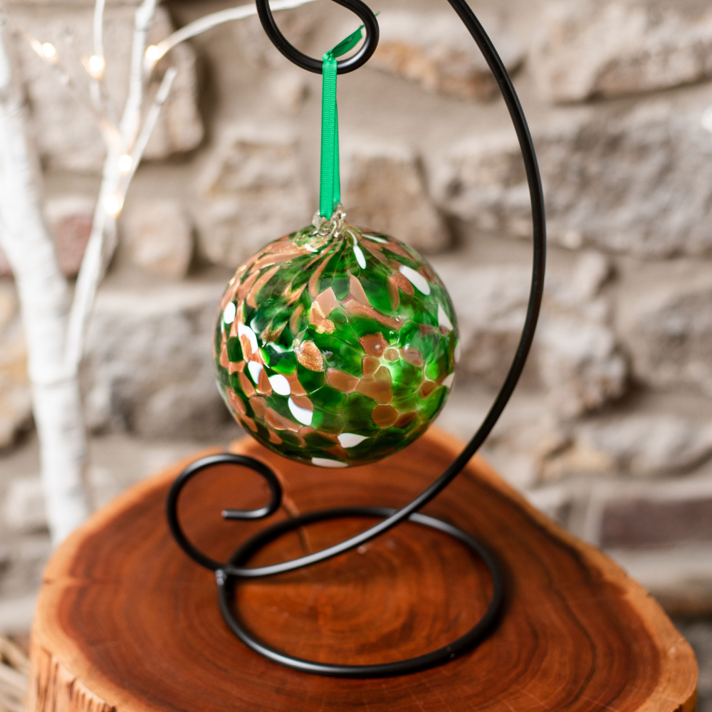Round glass ball with green, brown glitter and opaque white glass mosaiced around the entire surface on a black ornament holder on an end table. 