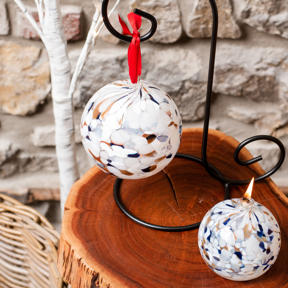 Round glass ball with opaque navy glitter, opaque copper glitter and opaque white glass mosaiced around the entire surface on a black ornament holder on an end table with a lit flat bottomed- round glass ball with a wick  with opaque navy glitter, opaque copper glitter and opaque white glass mosaiced around the entire surface. 
