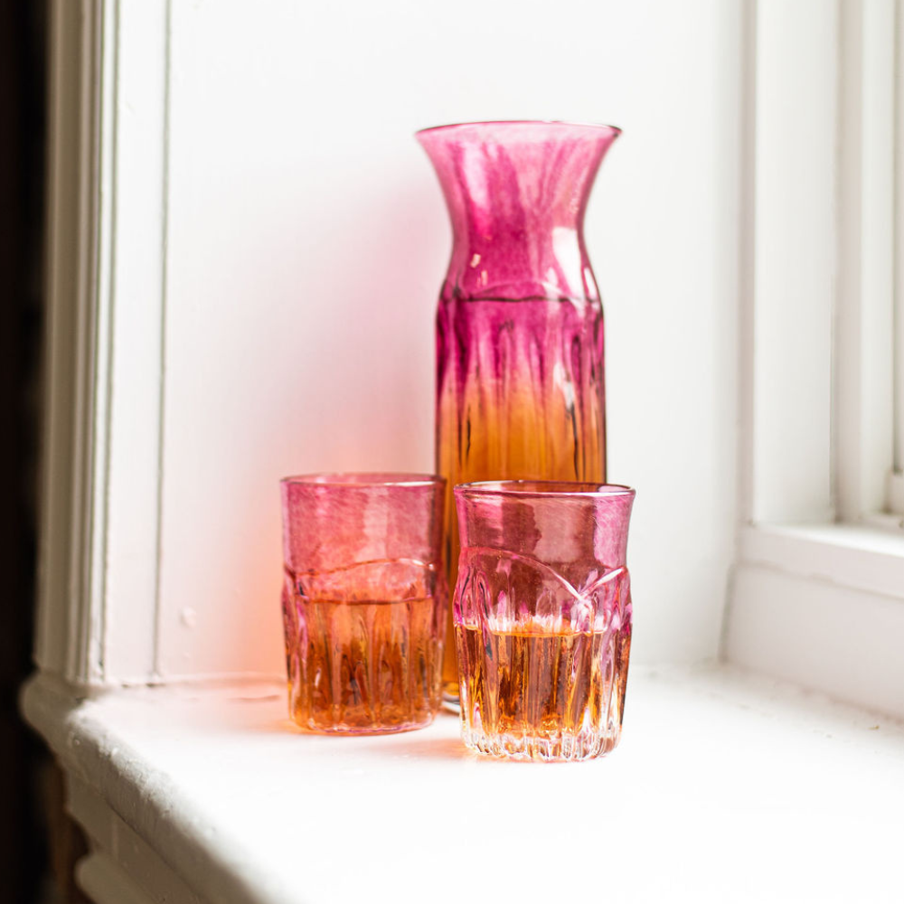 Carafe and two glasses with clear bottom and pink glass top, half filled with an amber liquid, on a white windowsill.