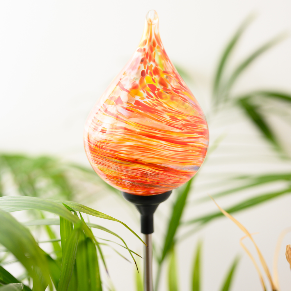 Tear drop glass orb with orange, red and amber glass swirled around the surface. There is a black clip at the base. Displayed amongst greenery.