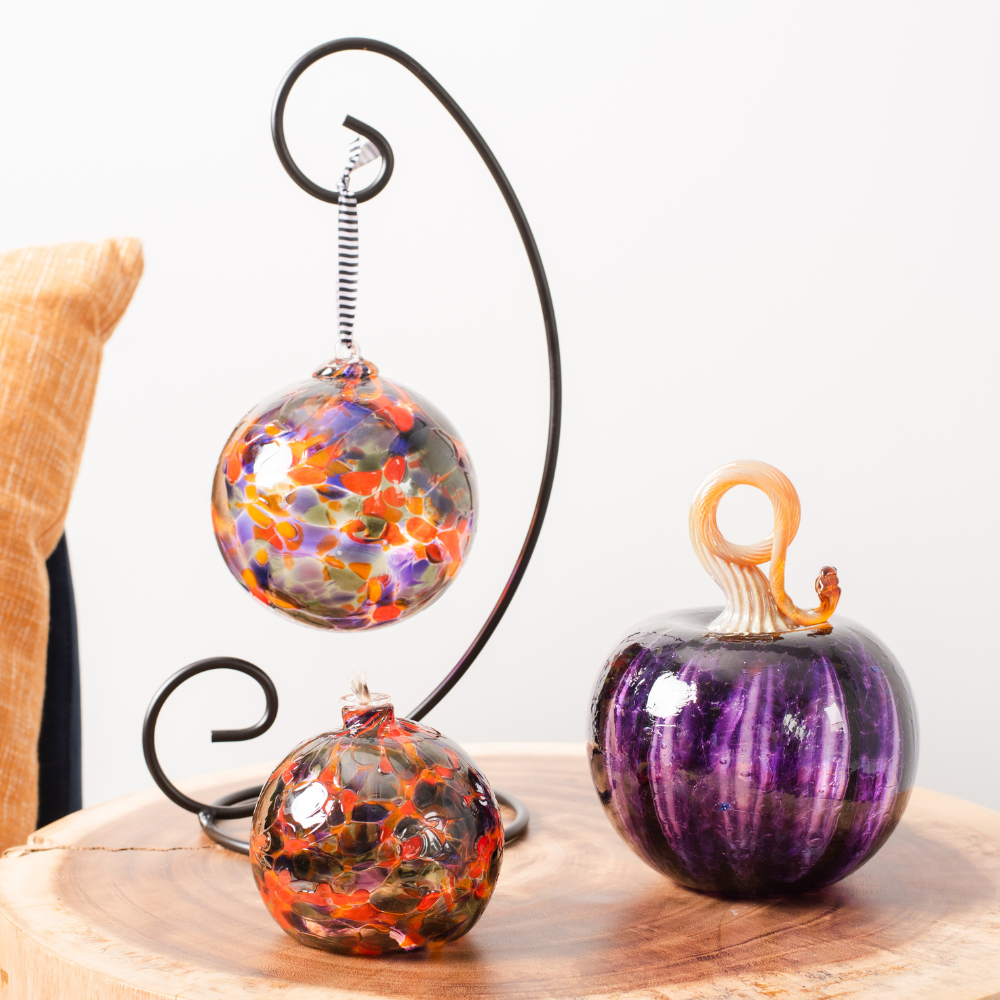 Round glass ball with purple, orange and grey glass mosaiced around the entire surface on a black ornament holder, flat-bottomed round glass ball with a wick with purple, orange and grey glass mosaiced around the entire surface and purple round pumpkin with a brown stem on an end table. 