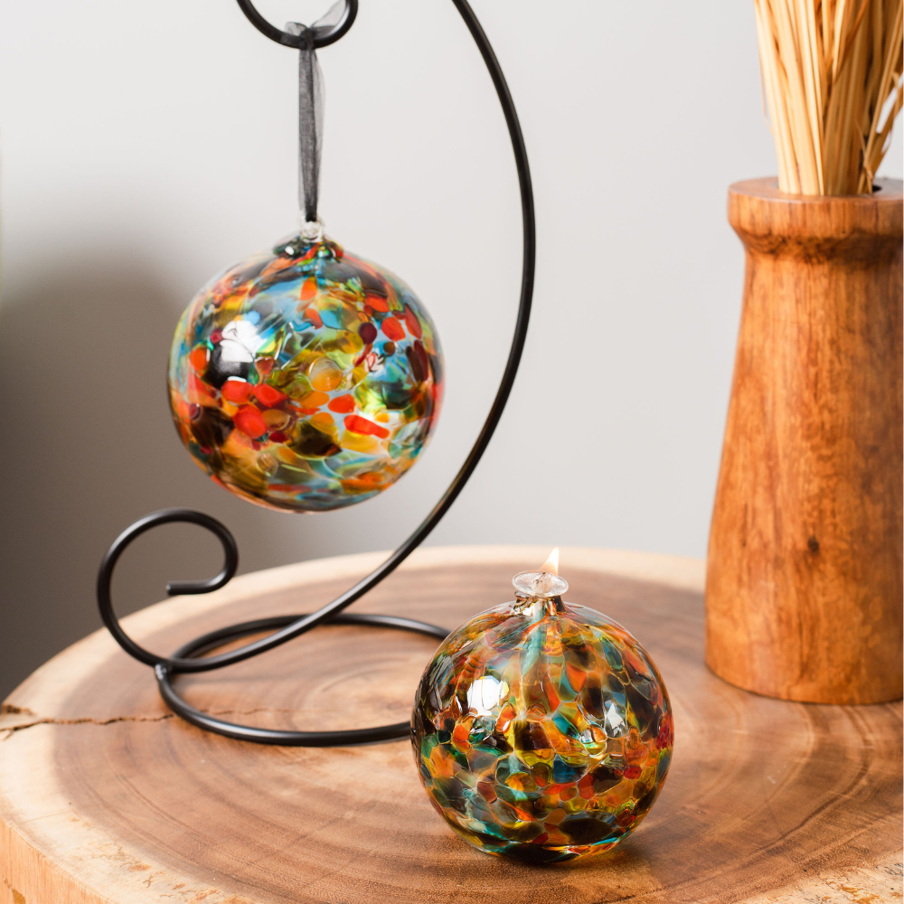 Lit Round glass ball with flat base and wick with brown, blue, red, lime, and orange glass mosaiced around the entire surface on a wooden table. On the left there's a black curved ornament holder with a round glass ball with brown, blue, red, lime, and orange glass mosaiced around the entire surface