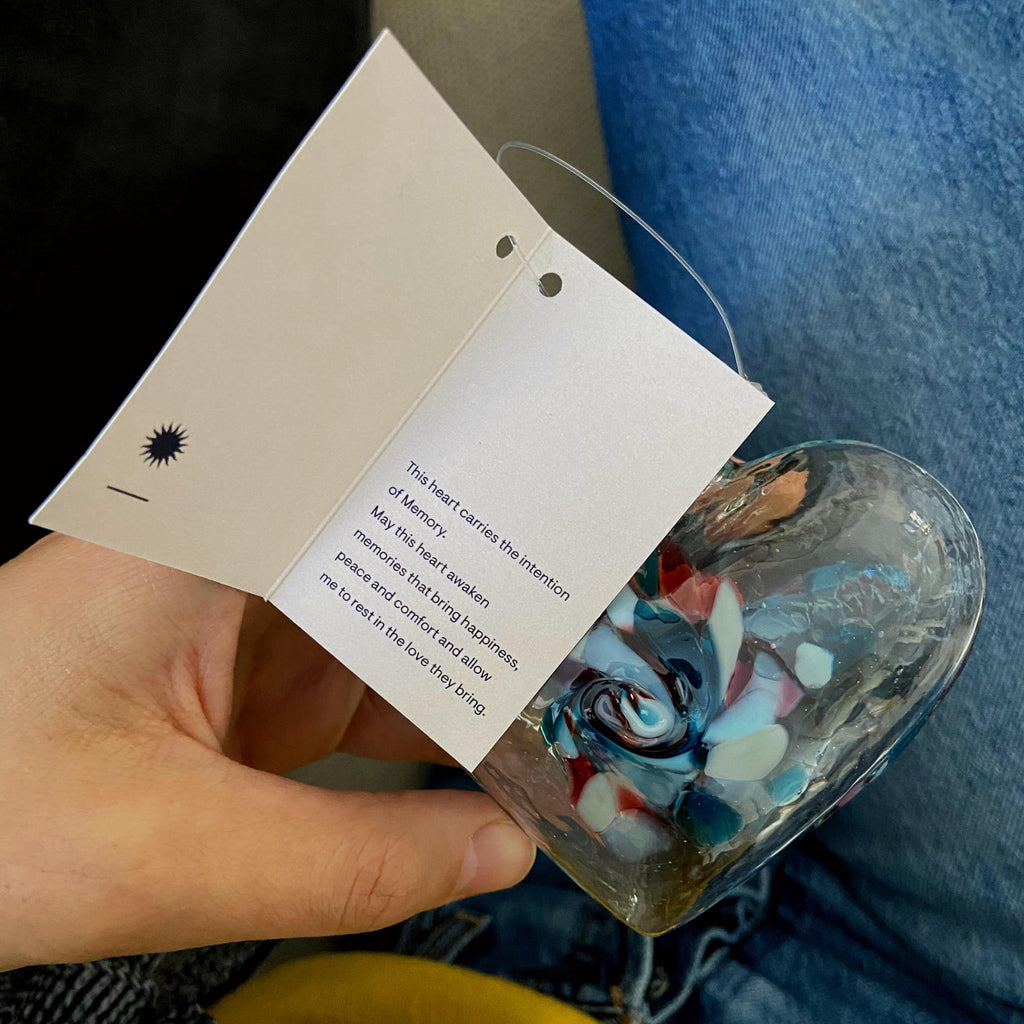 Clear glass heart with blue, pink, yellow and opaque white glass swirled in the centre resting in a hand above a jeaned leg. The Hear of Memories story card is open in the hand.
