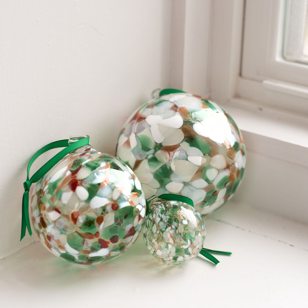 Three glass orbs of different sizes of Round glass ball with purple, orange and grey glass mosaiced around the entire surface. Each orb has a green ribbon and is sitting on a white window sill
