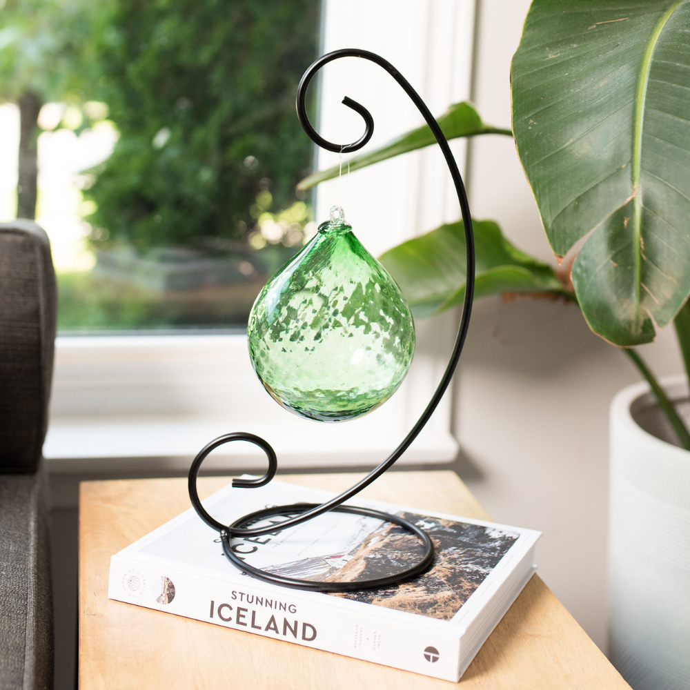 Green Verdant Vitality Radiance Orb on a Large Curved Single Ornament Holder resting on a book about Iceland on a wood end table. Green plant and window in the background