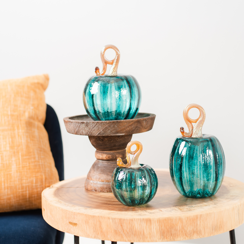 6" Teal Round Pumpkin on a wood pedestal. 3" Round and Oval Pumpkins on a wood table beside a blue chair with a yellow pillow