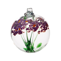 Clear glass orb with flat bottom and a wick with a green stem pulled from bottom to top to a canopy of purple opaque glass blossoms and orange centres on a transparent background