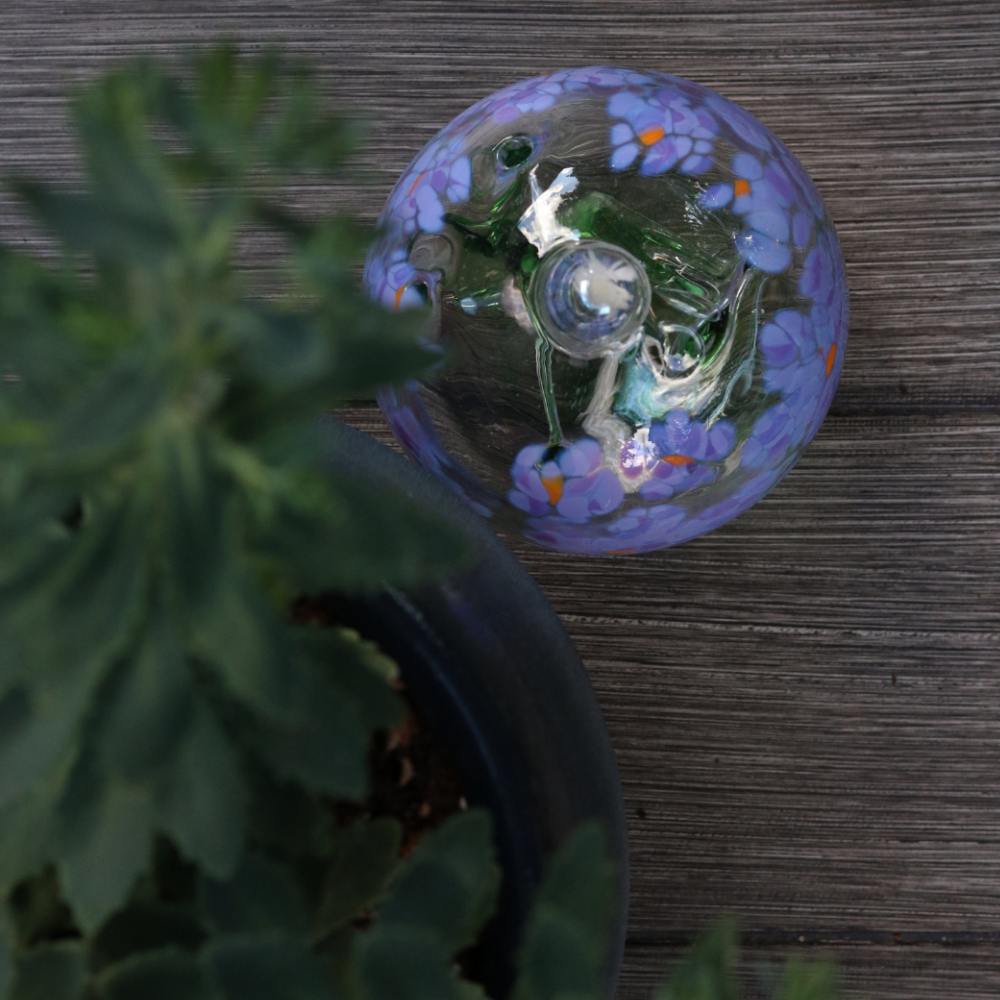 Clear glass orb with flat bottom and a wick with a green stem pulled from bottom to top to a canopy of purple opaque glass blossoms and orange centres on a wood table with greenery.