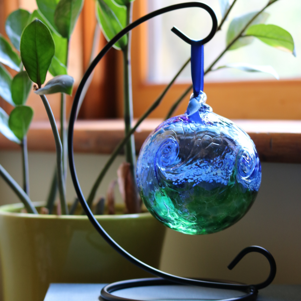 Glass orb with blue on top, white swirled across the middle, and green on the bottom hanging on a black curved ornament holder in front of a green plant and window.