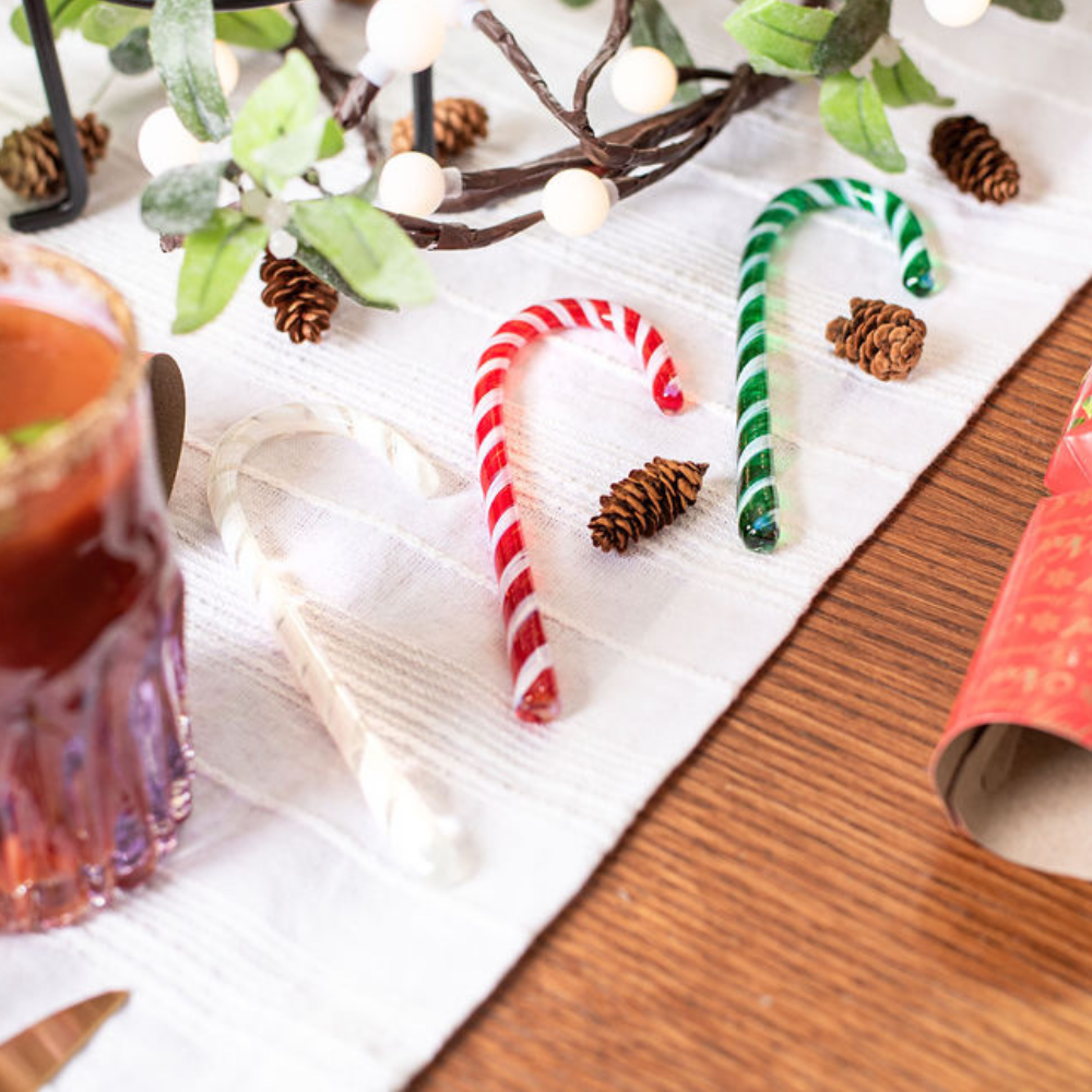 Red and white striped and Green and white striped glass Candy Canes resting on a white runner in a holiday table setting. Pinecones spread around. 