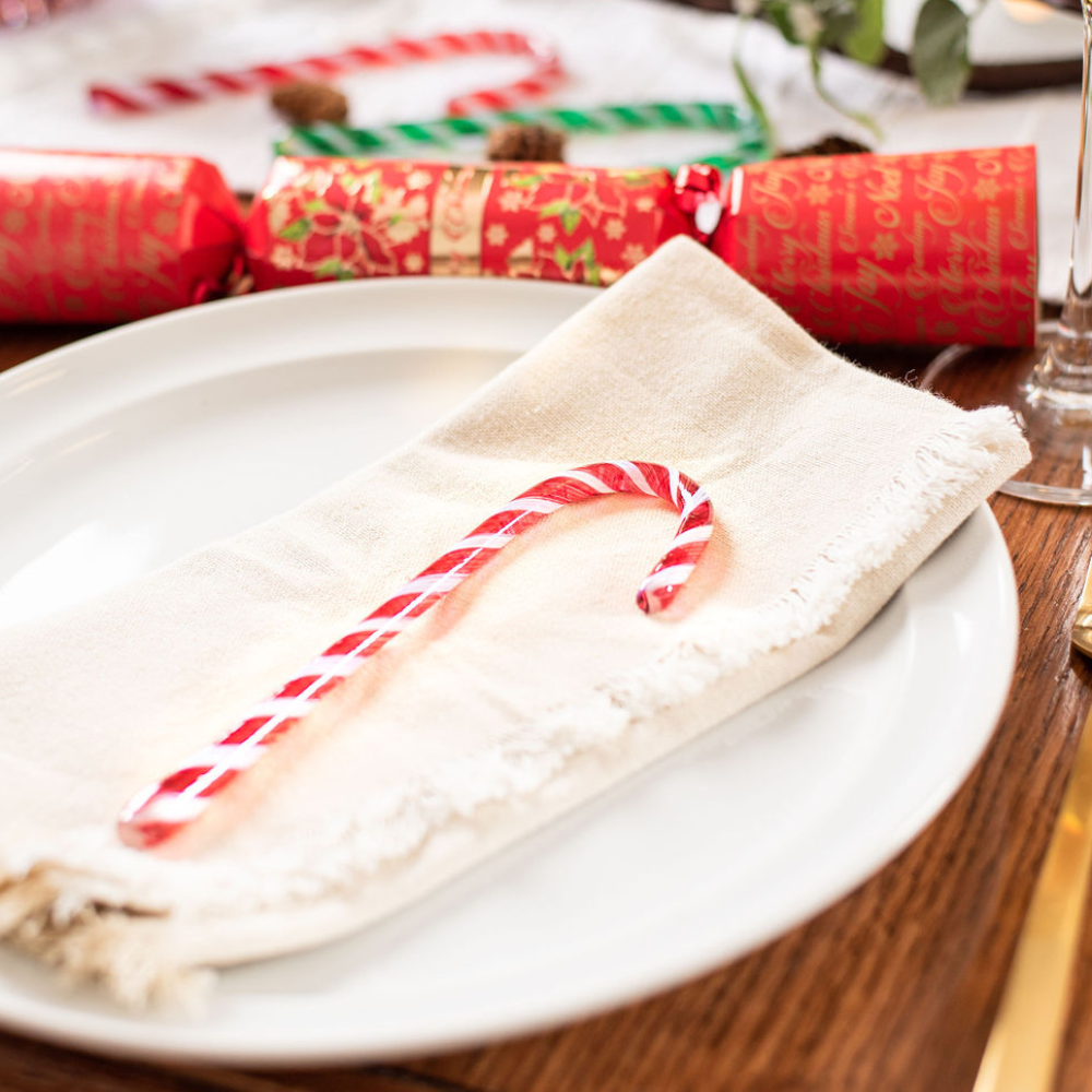 Red and white striped glass Candy Cane on a white napkin on a white place. A Christmas cracker is placed above. 