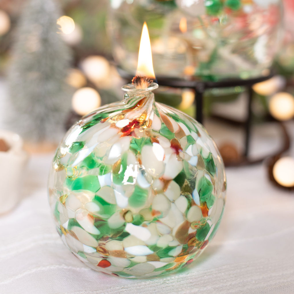 Lit Round glass ball with flat base and wick with green, red and opaque white glass mosaiced around the entire surface on a holiday table with a white table cloth