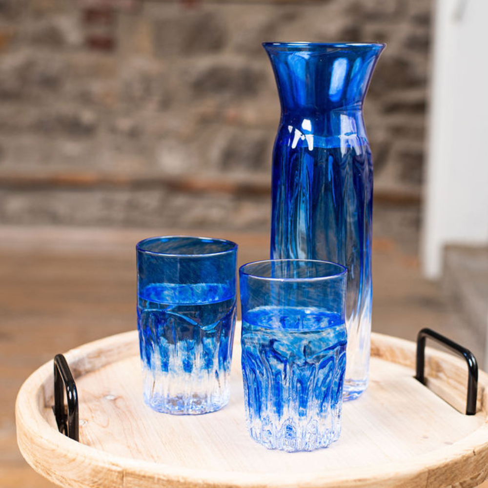 Carafe and two glasses with clear glass on the bottom and blue glass on the top. On a wood tray with a stone wall in the background.