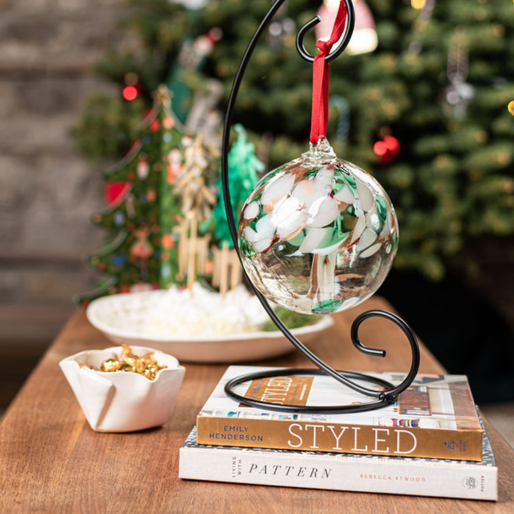 Large Curved Single Ornament Holder with a 6" Tree of Christmas. Holder is resting on a book that's on a coffee table. A Christmas Tree is in the background. 
