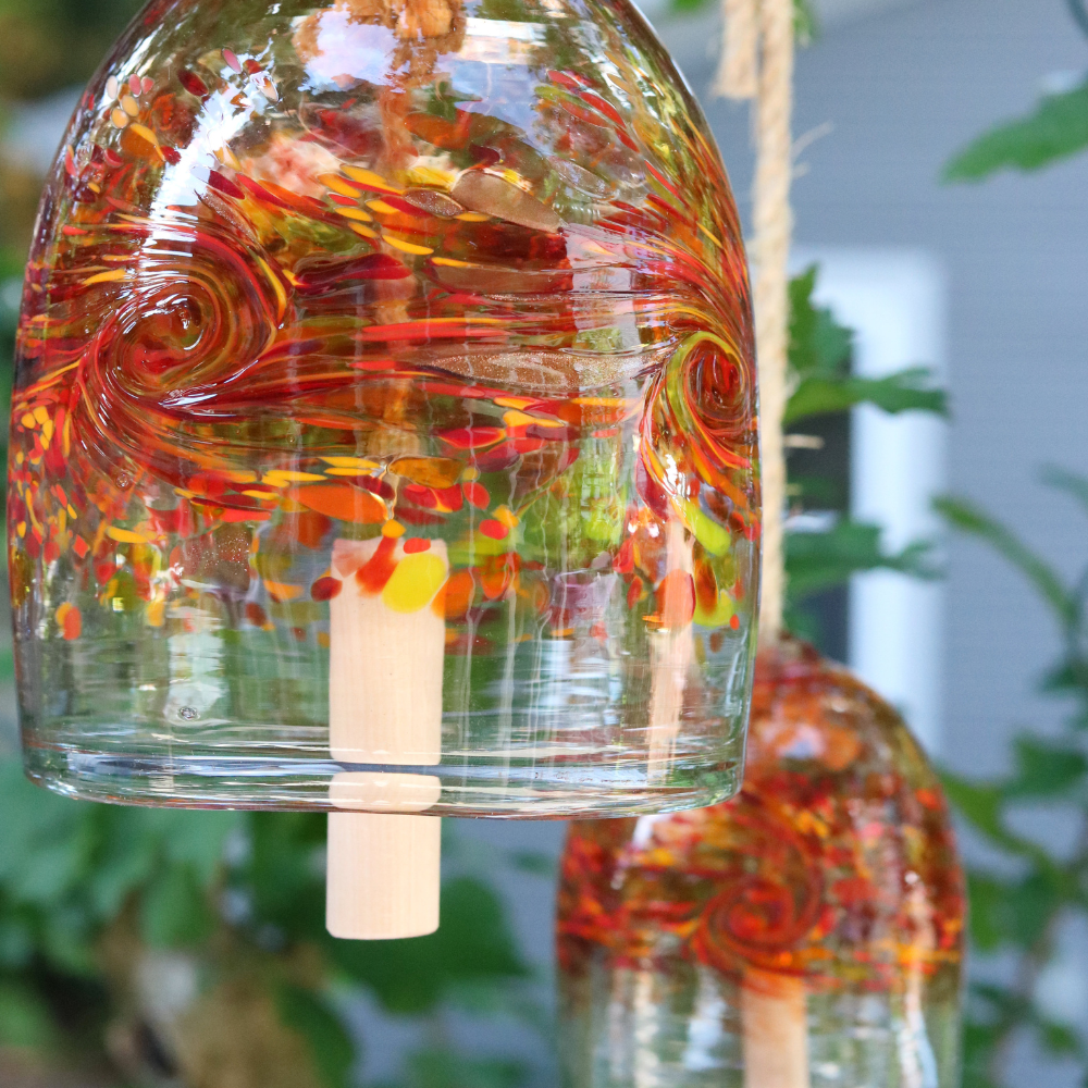 A clear glass bell with red, orange, yellow and purple glass swirled across the middle. A wooden toggle and rope are pulled through the middle. Hanging amongst greenery.