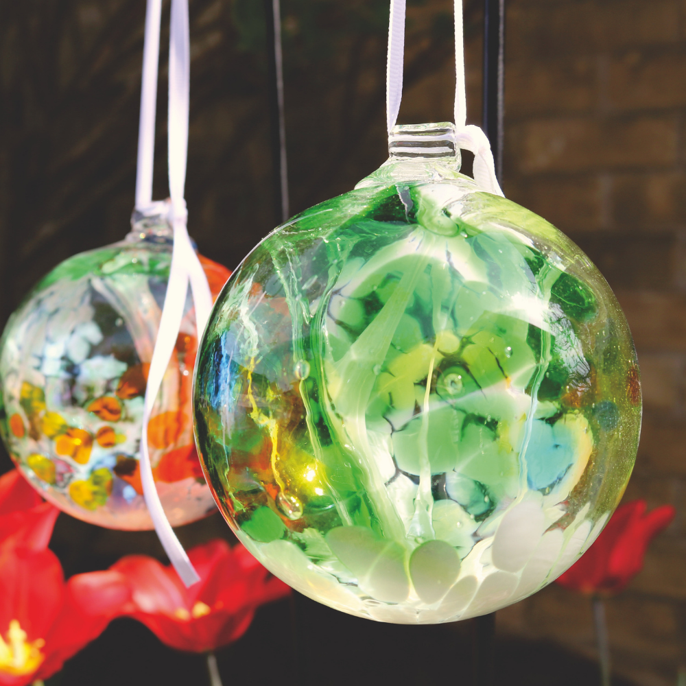 Glass orb with opaque white, green, orange, yellow and blue glass on the surface with white webbing pulled from bottom to top hanging on a white ribbon in front of another glass orb with orange, white, yellow and green glass on the surface with white webbing pulled from bottom to top amongst red flowers.