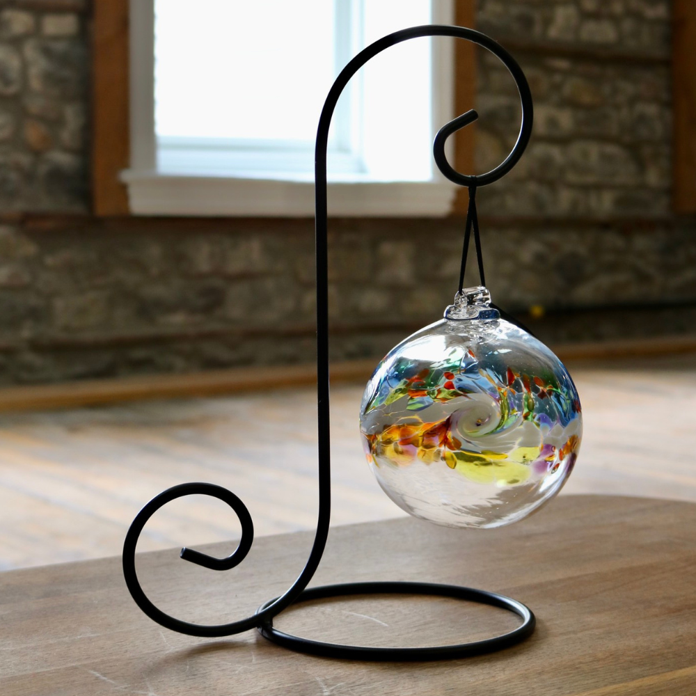 Glass orb with a colourful band across the middle. Blue, orange and green glass on top, white glass swirled in the middle and yellow, orange and red glass swirled below  on a black ornament holder in front of a window.