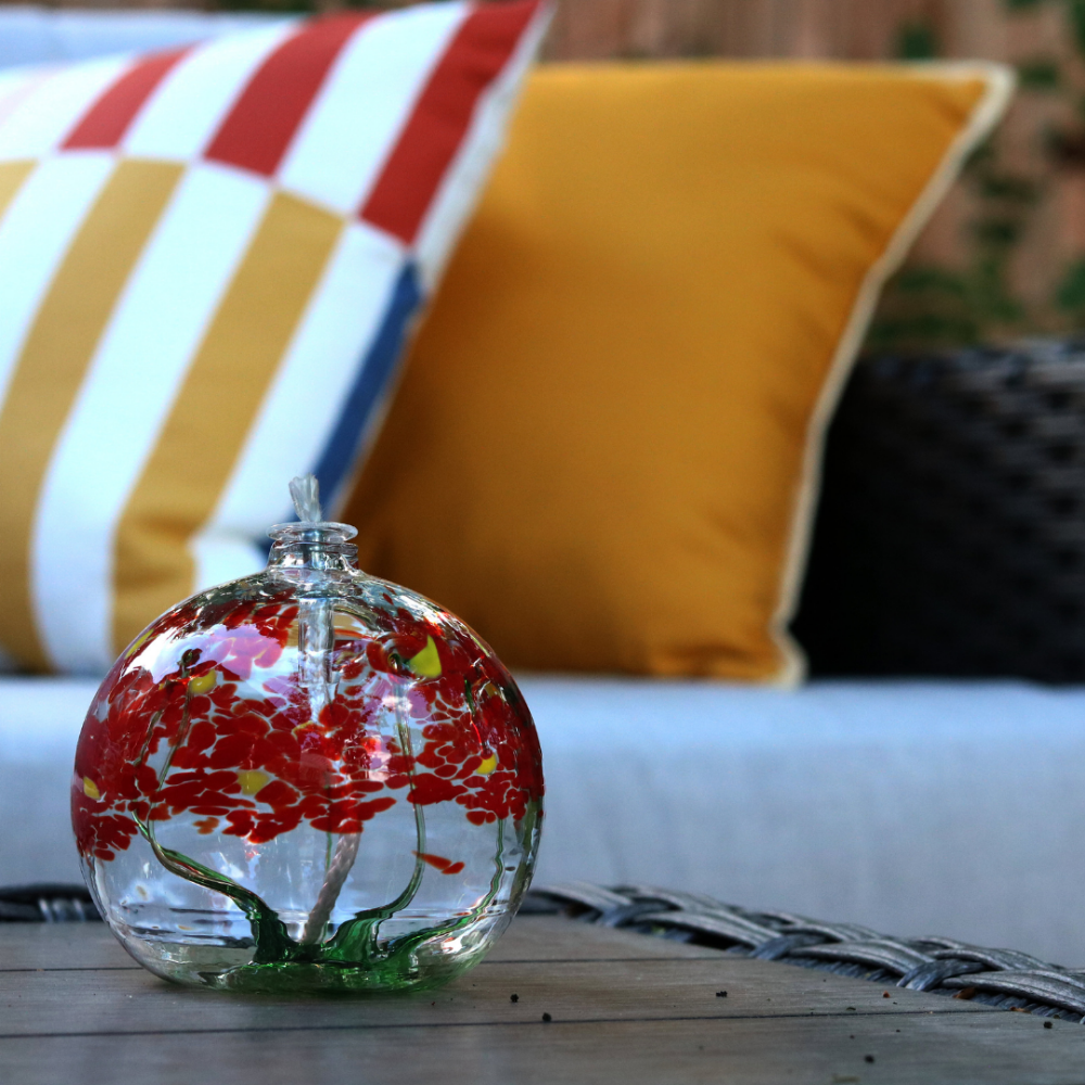 Clear glass orb with flat bottom and a wick with a green stem pulled from bottom to top to a canopy of red opaque glass blossoms and yellow centres on a wood table with patio furniture in the background.