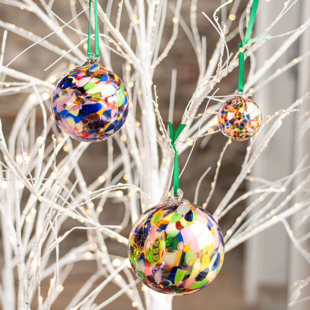 Three different sized round glass balls with opaque pink, blue, yellow, purple, opaque green, opaque yellow, lime and opaque white glass mosaiced around the entire surface hanging from green ribbons on a white tree with twinkle lights. 