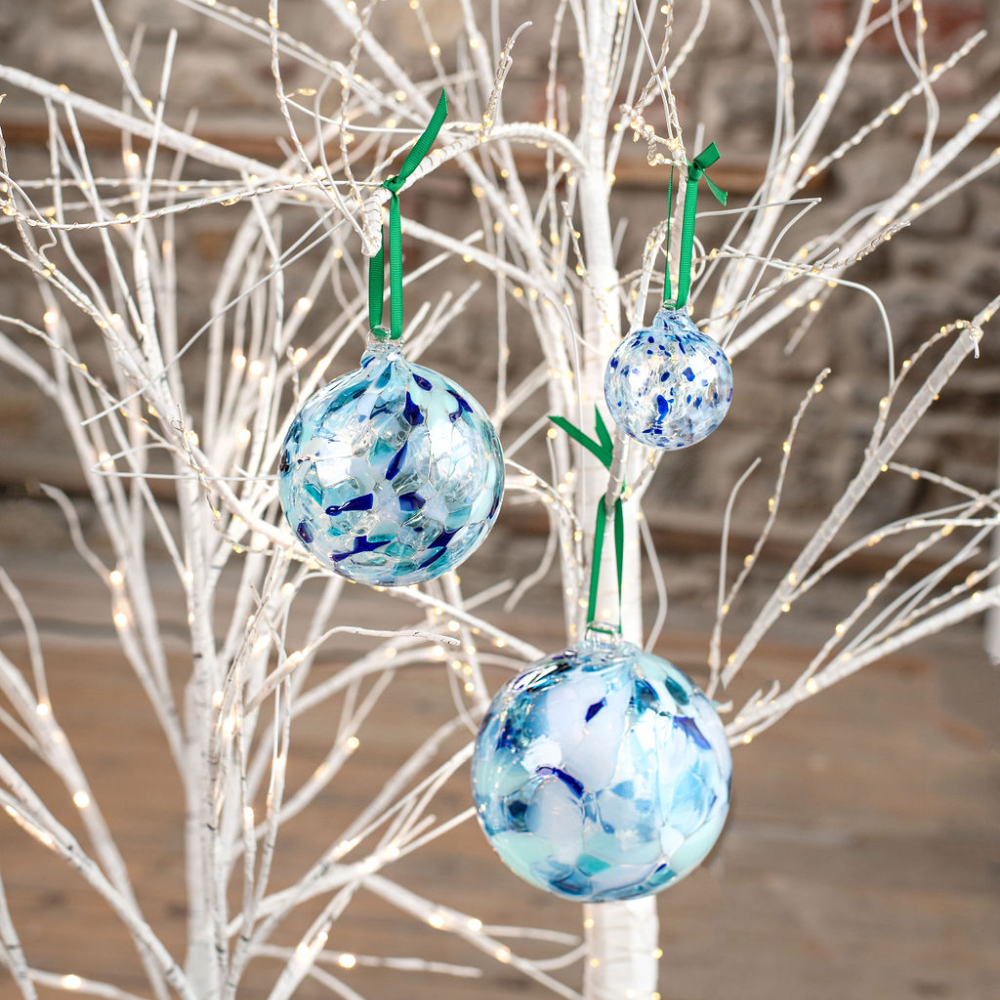 Three Round glass balls with opaque blue, light blue and opaque white glass mosaiced around the entire surface hanging by green ribbon on a white tree with twinkle lights. 