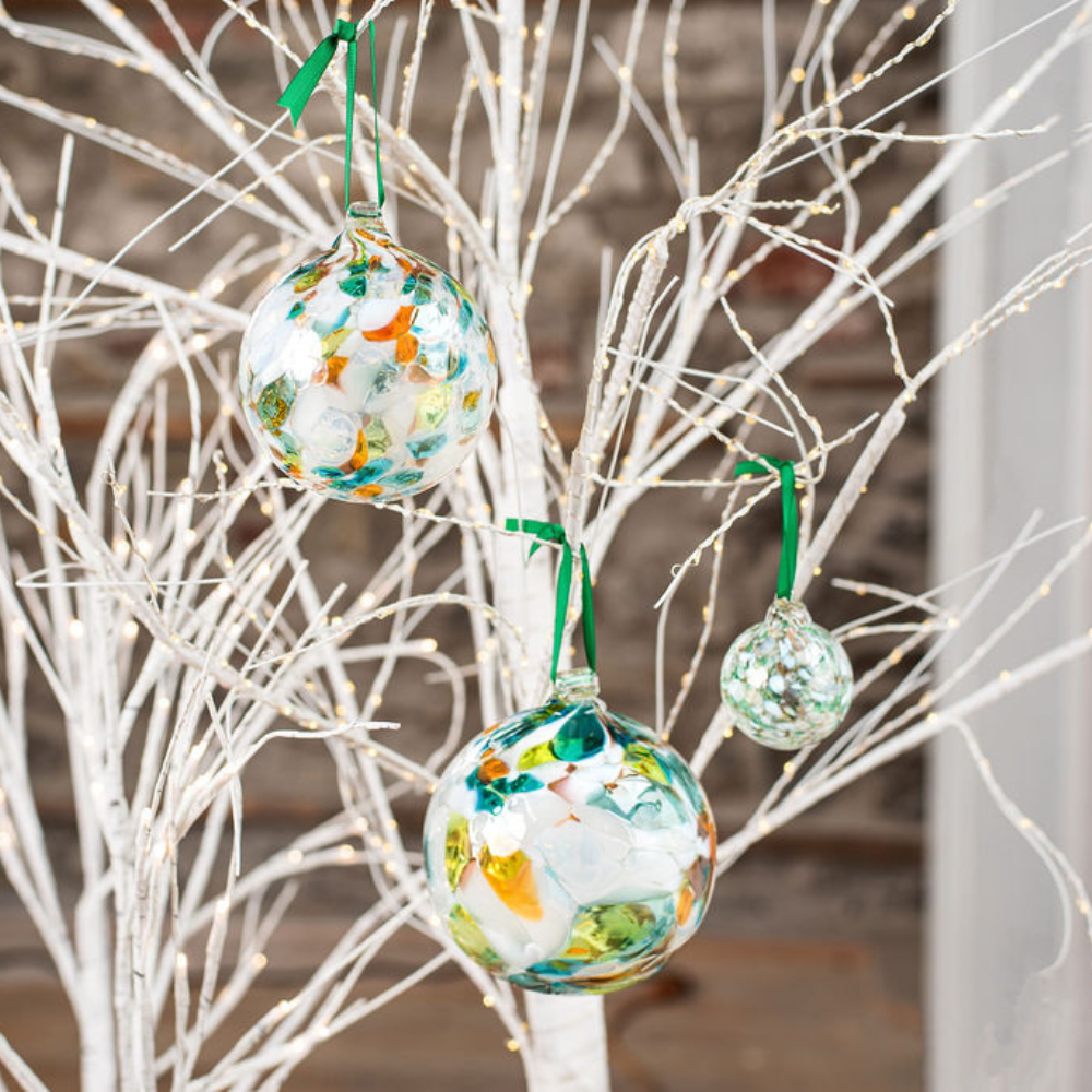 Three different sized glass balls that have Lime, orange, teal and opaque white glass mosaiced around the entire surface displayed on a white tree, hanging with a green ribbon