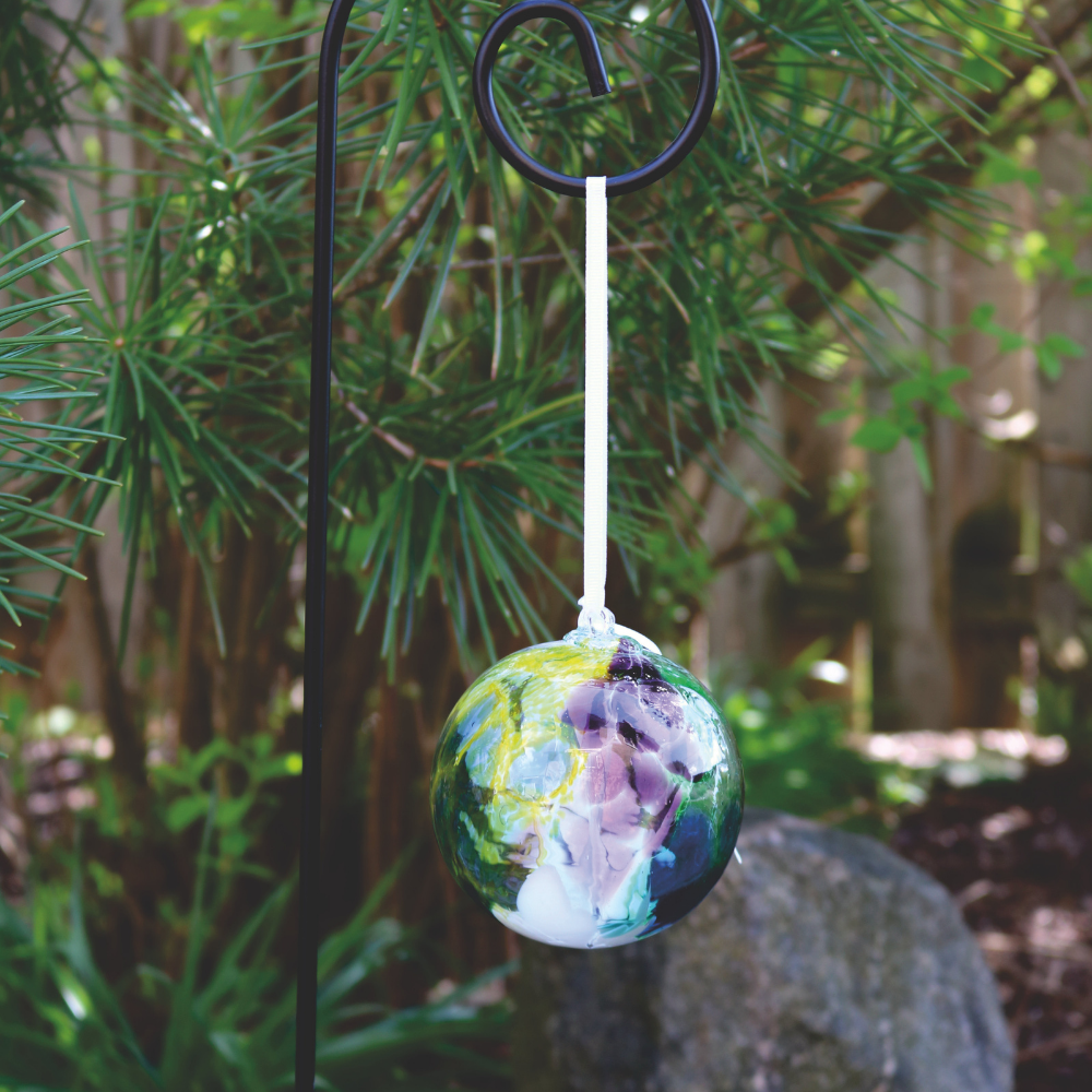 Glass orb with opaque white, blue, lime and purple glass on the surface with white webbing pulled from bottom to top hanging on a white ribbon from on a black shepherd's hook stake in a garden.