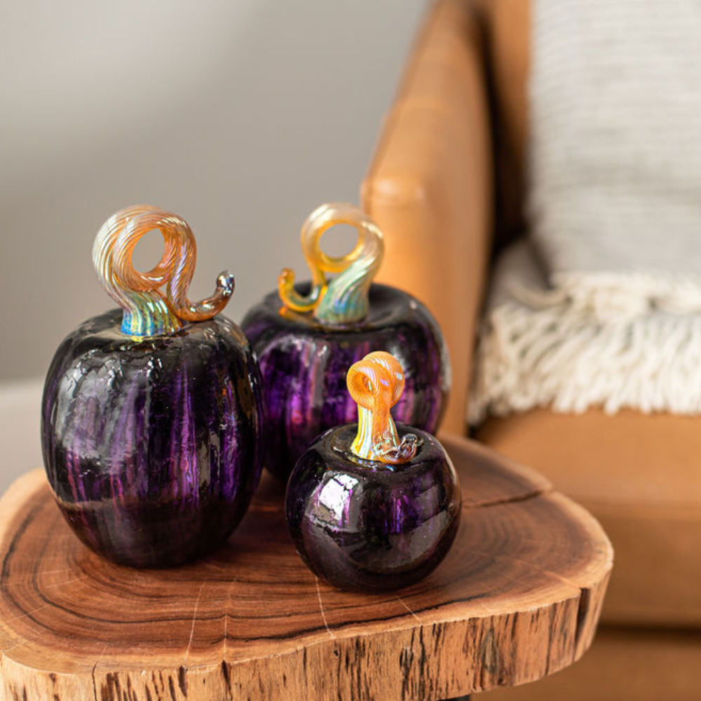 Oval, 3" and 6" Round Purple Pumpkins on a wooden table with a brown chair in the background.