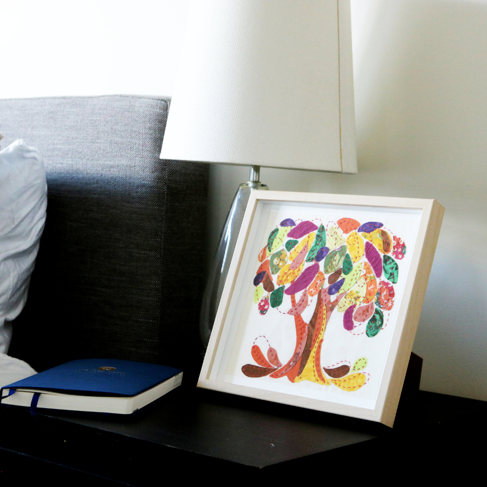 orange, brown, cranberry, yellow, green and blue fabric tree on a white background in a wood frame. Displayed on a black side table beside a blue notebook