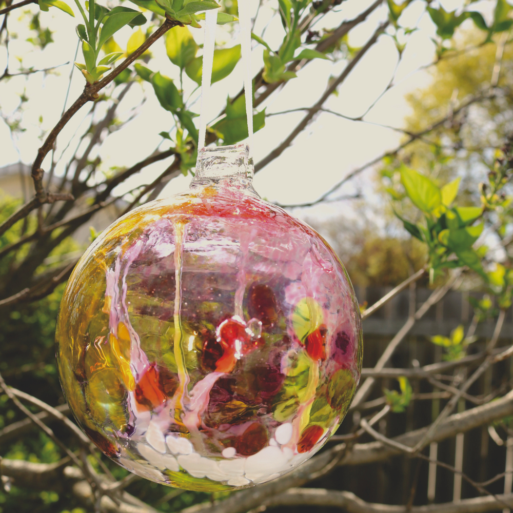 Glass orb with opaque white, pink, lime and orange glass on the surface with white webbing pulled from bottom to top hanging on a white ribbon from a tree branch.