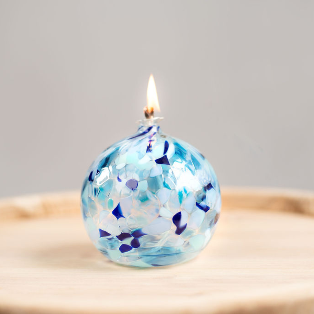 Lit Round glass ball with flat base and wick with opaque blue, light blue and opaque white glass mosaiced around the entire surface on a wooden tray