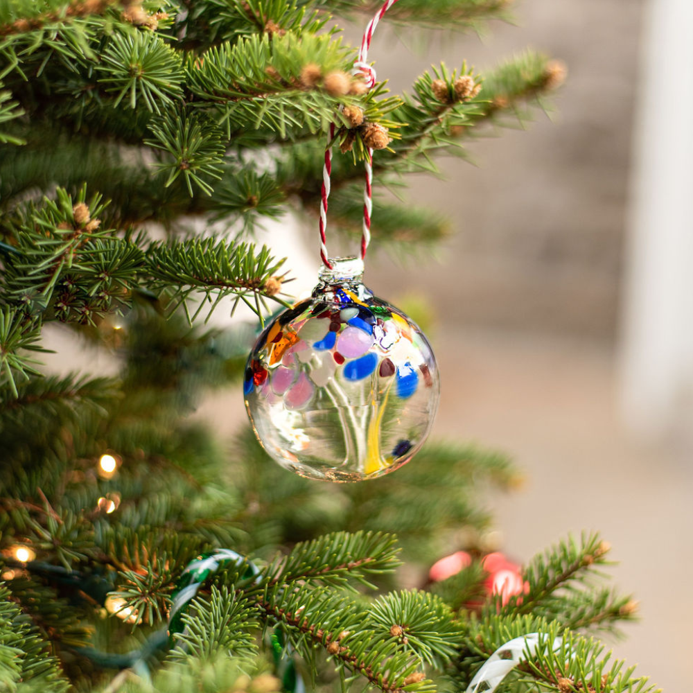 Clear glass orb with orange glass pulled through the middle and Opaque White, Pastel Pink, Amethyst, Cobalt, Amber, Orange, Opaque Red, Green and Brown bits on top hanging on a green Christmas Tree with a red and white string