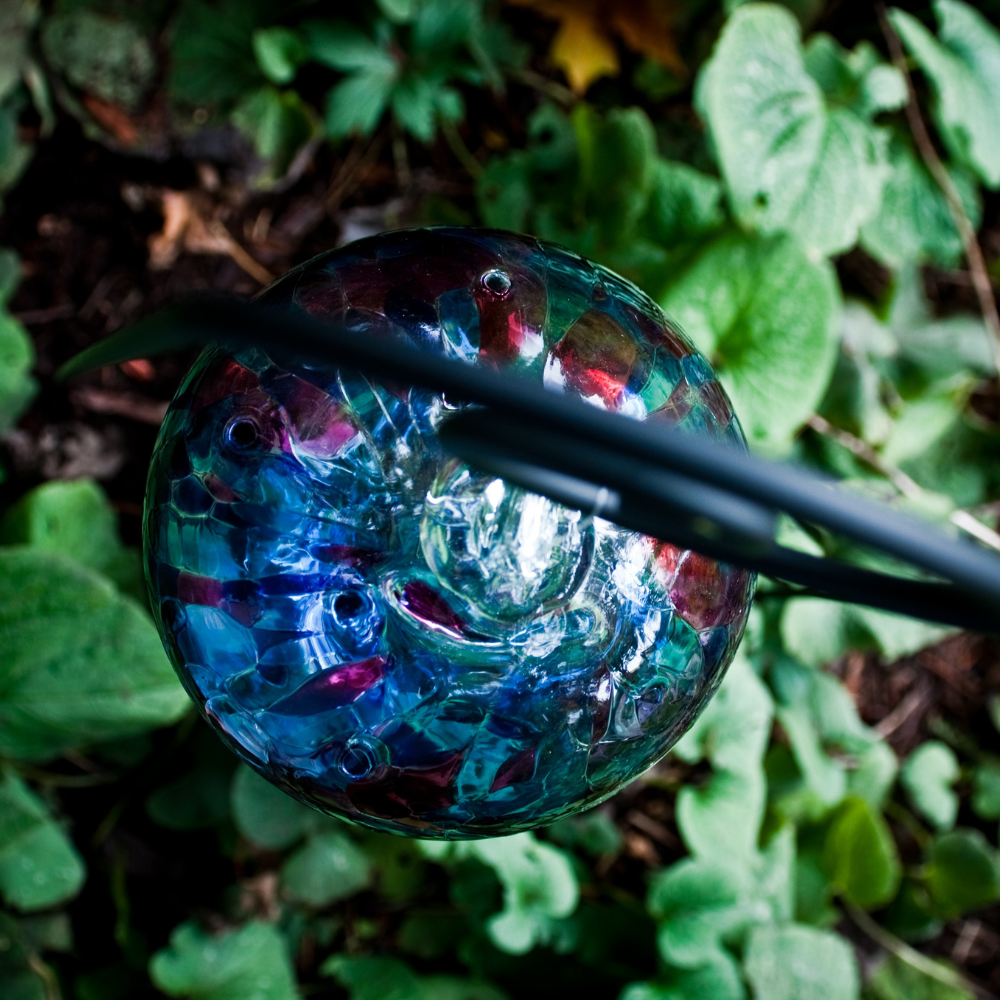 Clear glass orb with amethyst and purple glass pulled through the middle and Blue, Dark Blue, Amethyst, Purple and Red bits on top on a black Shepherd's Hook Stake amongst greenery
