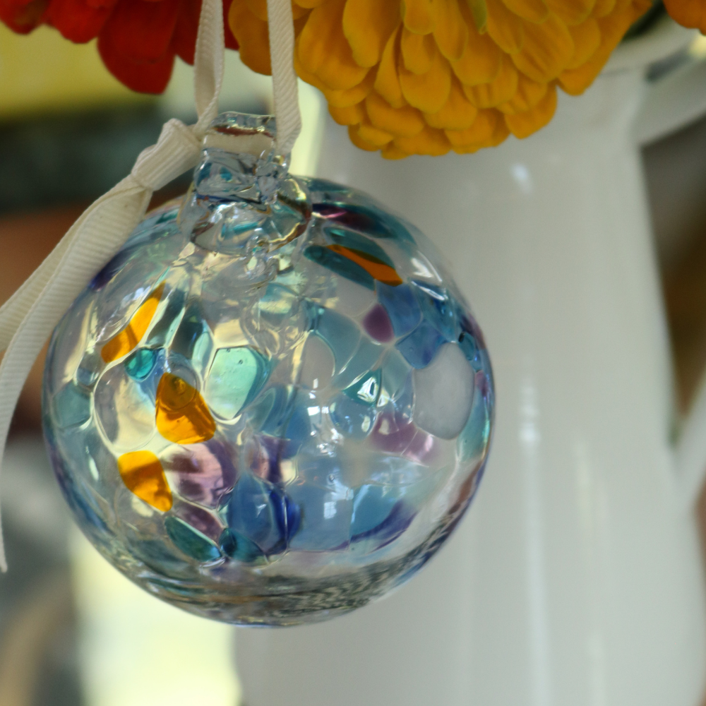 Clear glass orb with pink glass pulled through the middle and opaque white, blue, purple and orange bits hanging on a white ribbon with orange and red flowers in a white vase in the background