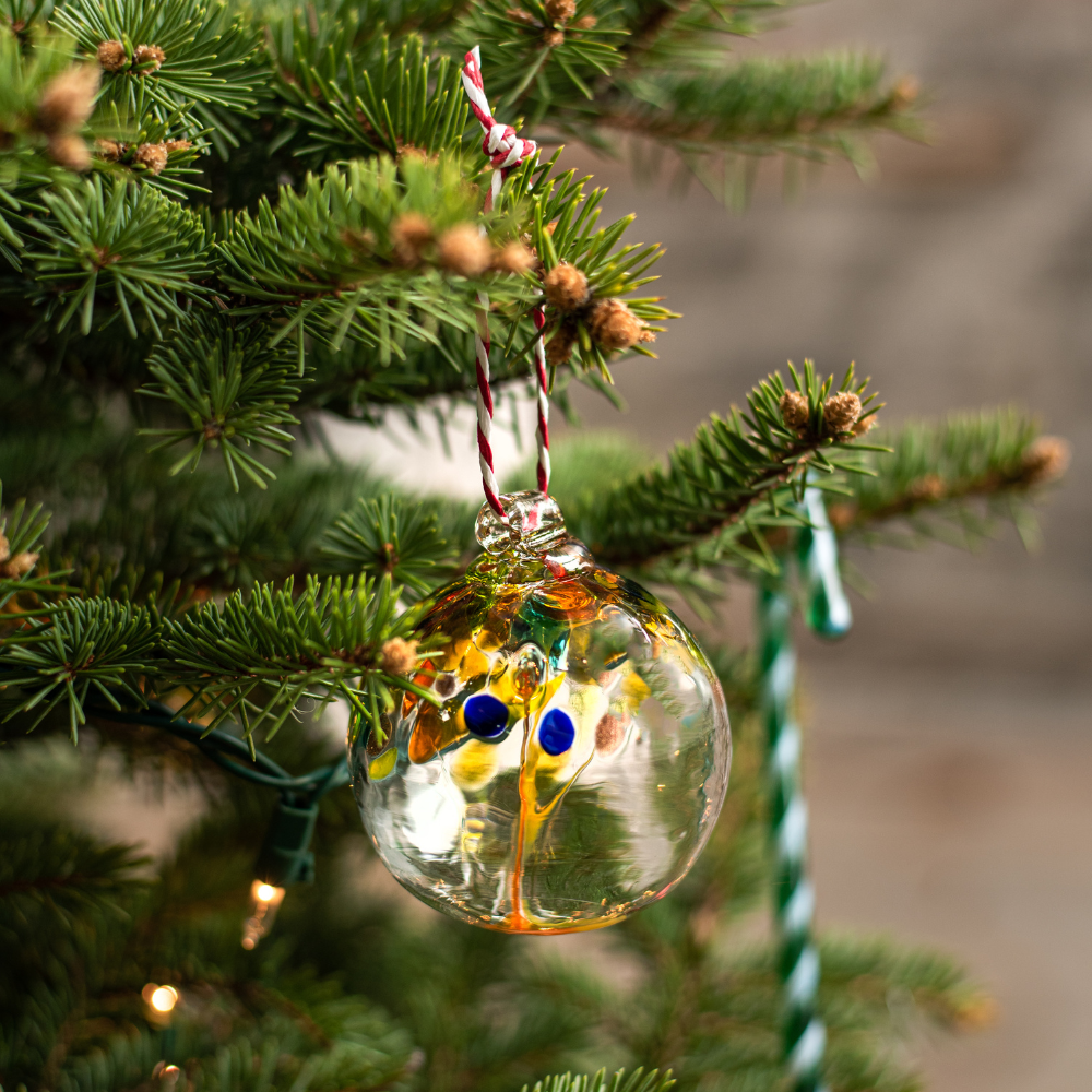 Clear glass orb with clear, orange and blue glass pulled through the middle and yellow, red, orange, gold glitter and blue bits on top hanging on a green Christmas Tree with a green glass Candy Cane in the background