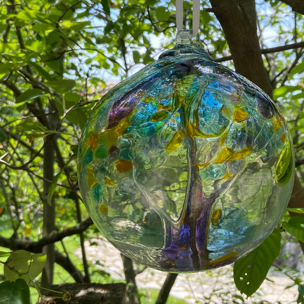 Clear glass orb with clear and purple glass pulled through the middle and yellow, purple, pink, orange, green and blue bits on top hanging from a tree