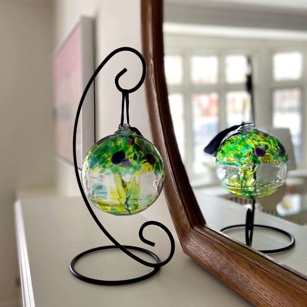 Clear glass orb with purple and green glass pulled through the middle and yGreen, Teal, Lime, Blue, Purple and Pink bits on top on a black ornament holder. Displayed on a white mantel in front of a mirror. Can see reflection of the green orb in the mirror. 