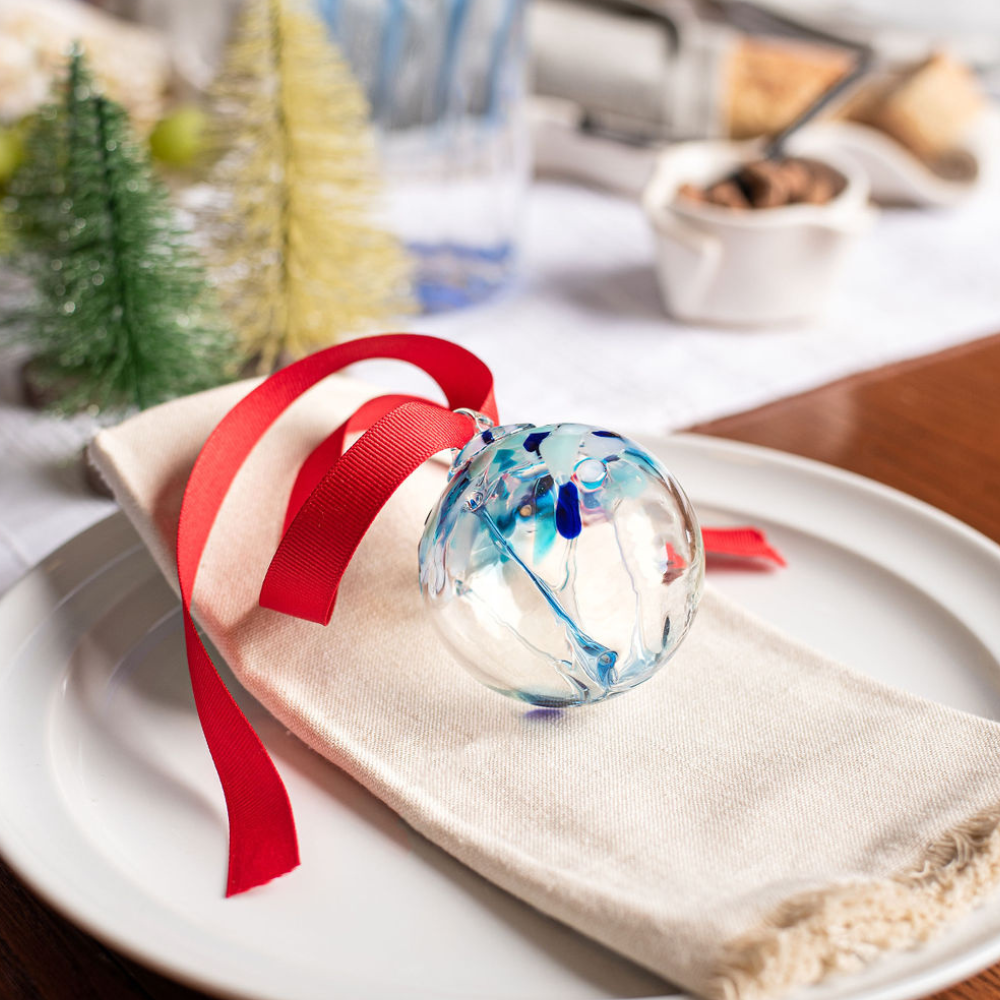 Clear glass orb with blue glass pulled through the middle and white and blue bits on top with a red ribbon, resting on a white napkin on a white table setting. Small Christmas Trees in the background. 