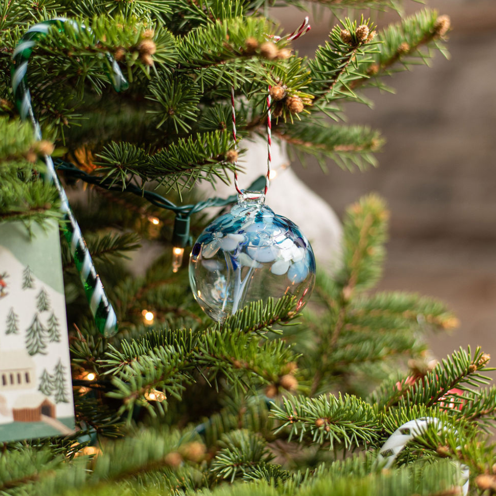 Clear glass orb with blue glass pulled through the middle and white and blue bits on top hanging with a red and white string from a Christmas Tree. 