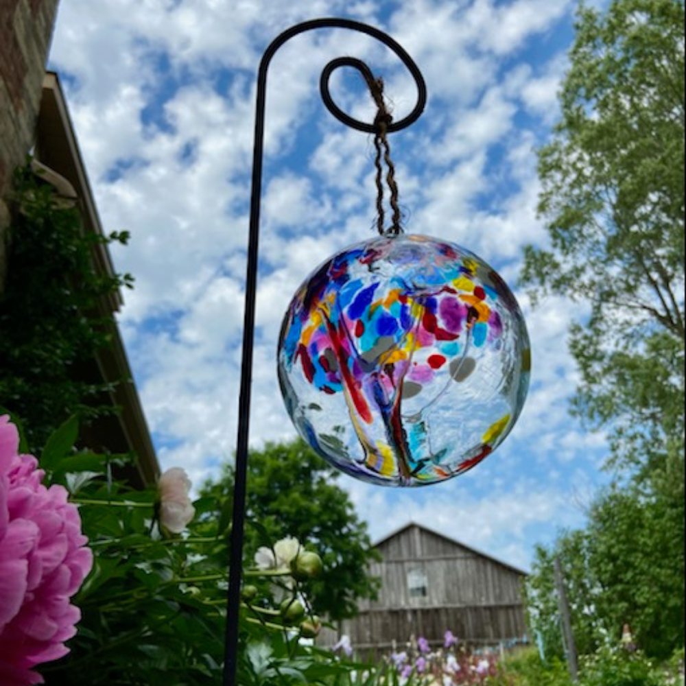 Clear glass orb with purple, orange, yellow and blue glass pulled through the middle and yellow, white, red, purple, pink, orange and blue bits on top on a black Shepherd's Hook Stake in a garden. Blue sky with clouds in the background. 