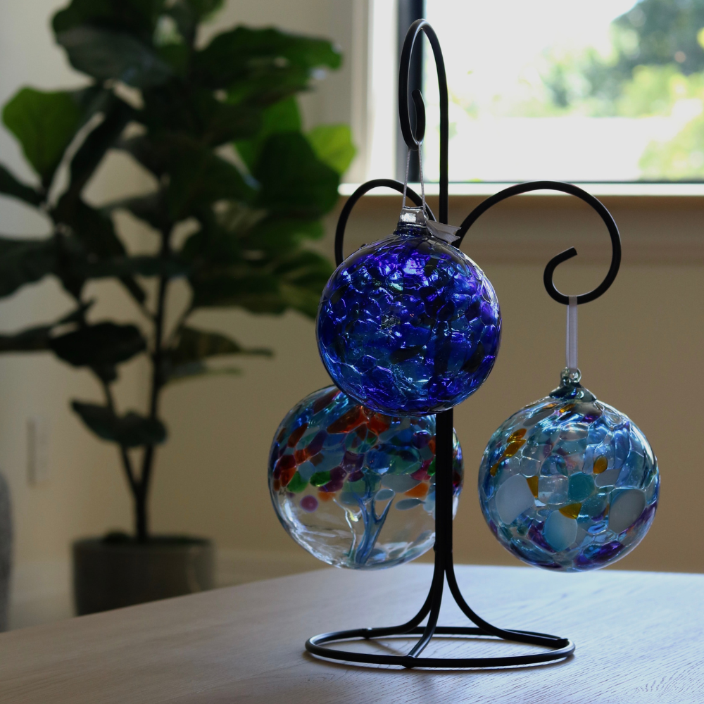 6" Winter Solstice Calico Ball, 6" Stormy Sea Calico Ball and 6" Tree of Caring on a Triple Ornament Holder. Displayed on a table with a tree and window in the background.
