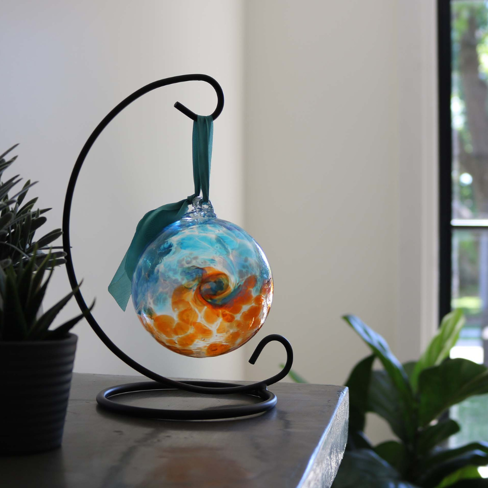 Glass orb with aqua on top, white swirled across the middle, and orange on the bottom hanging on a black curved ornament holder beside a green plant and window.